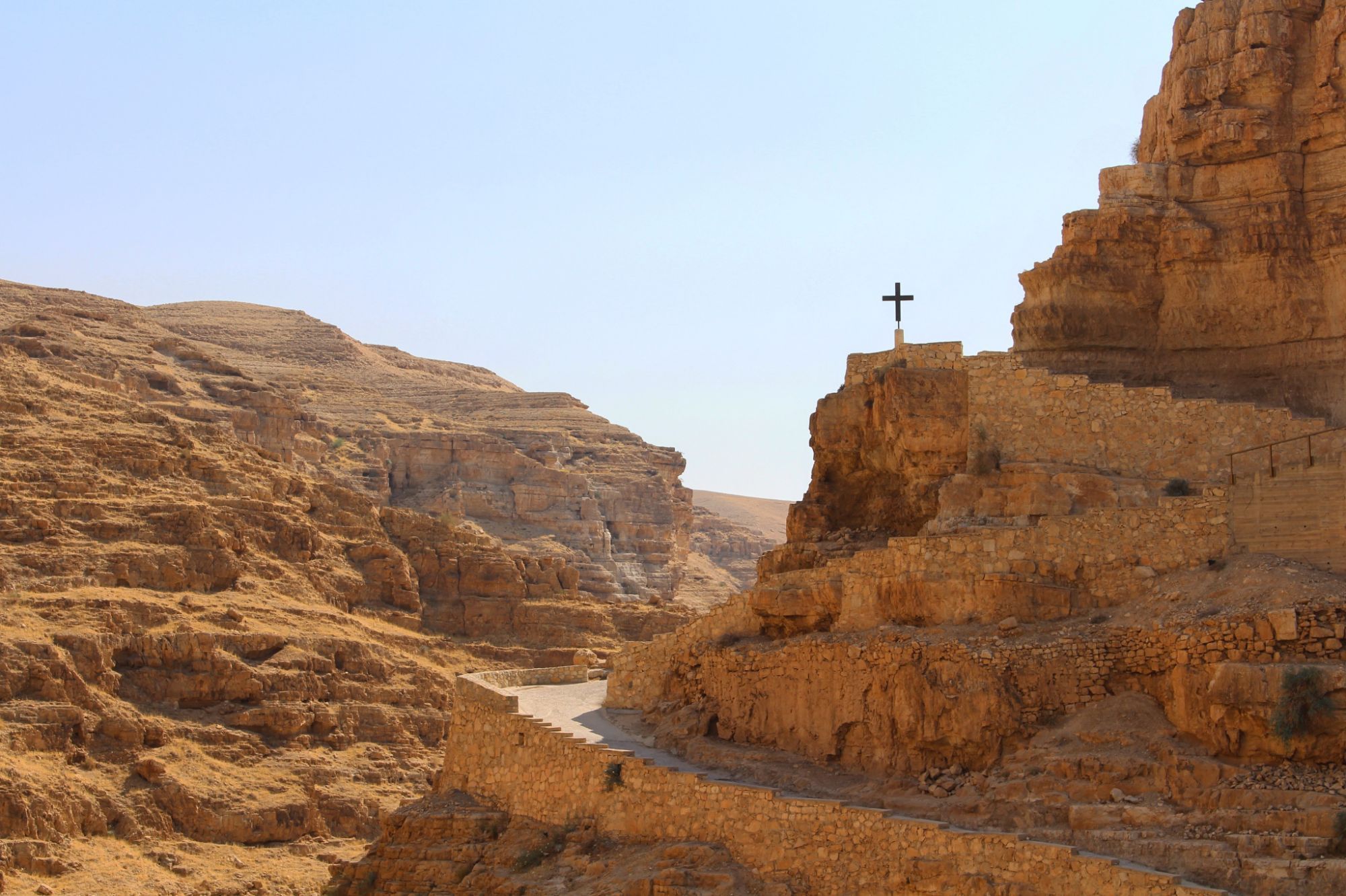A monastery built into the cliffs of a desert canyon, a cross standing above its walls